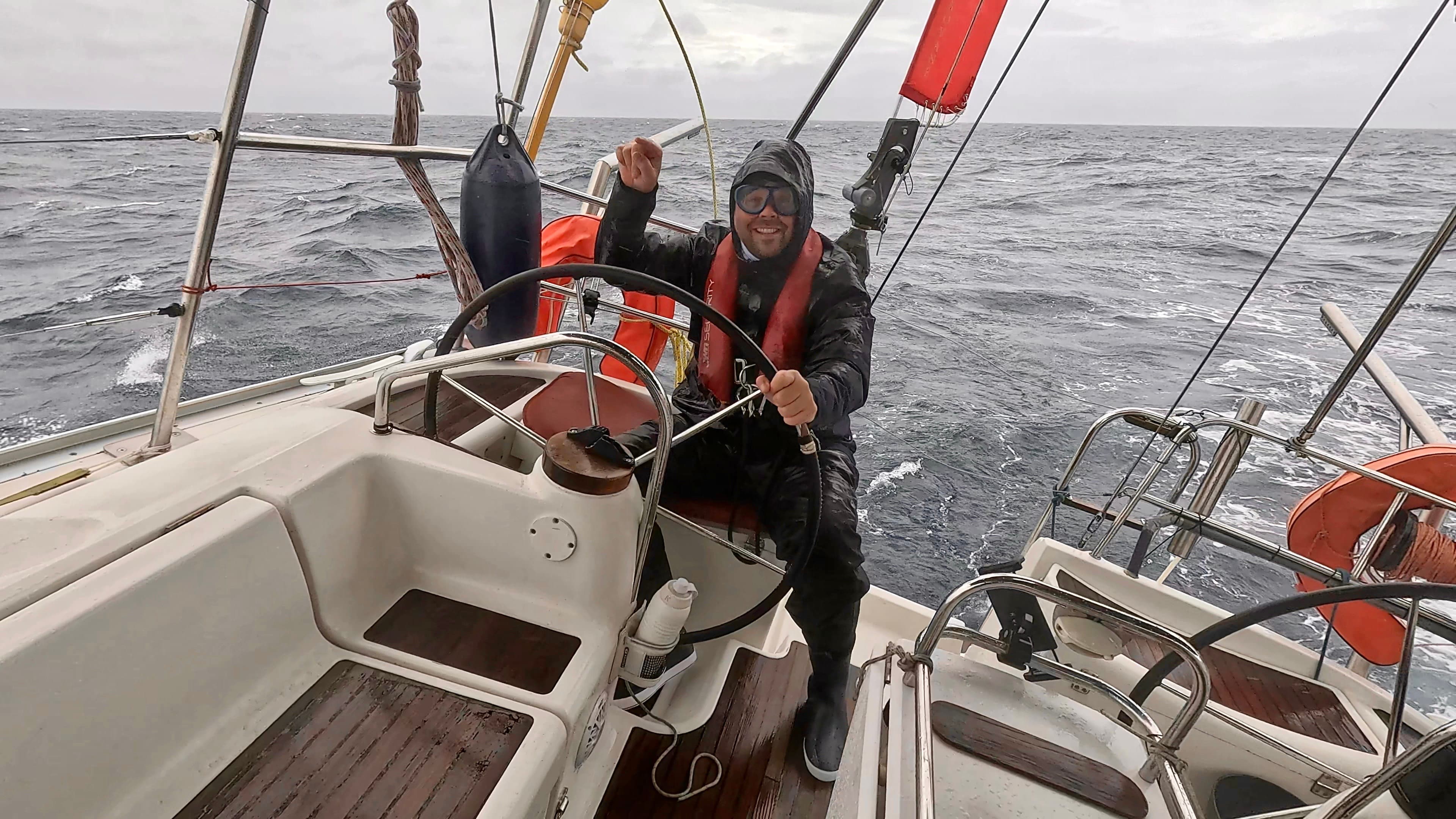 Ein Mann steuert die Segelyacht Rocinante, während der Atlantiküberquerung 2026, im Sitzen in voller Segelbekleidung und